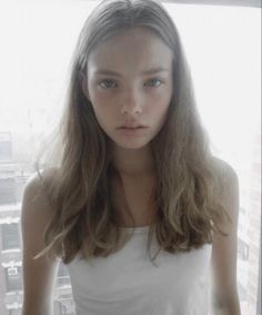 a young woman with long hair and blue eyes looking at the camera while standing in front of a window