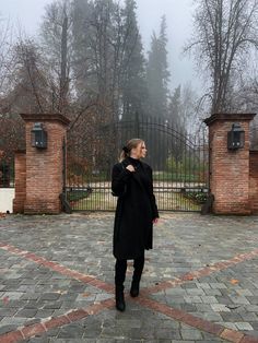 a woman standing in front of an iron gate on a cobblestone walkway with trees and bushes behind her