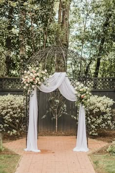 an outdoor wedding ceremony with white drapes and flowers on the arch, surrounded by greenery