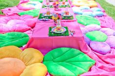 a long table covered in pink, green and yellow pillows on top of grass next to a field