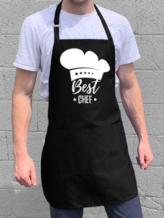 a man wearing an apron with the words best chef in white on it, standing next to a brick wall