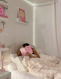 a woman laying in bed reading a pink book with lights on the wall behind her