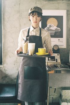 a woman in an apron holding a tray with two cups on it and a coffee maker behind her