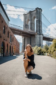 a woman squatting down in front of the brooklyn bridge, looking at the camera