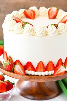 a white cake topped with strawberries on top of a wooden stand next to a bowl of strawberries