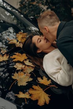 a man and woman kissing in front of a car with autumn leaves on the hood