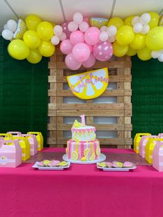 a pink table topped with a cake and lots of yellow and pink balloons above it