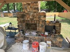 Construction zone of a DIY outdoor fireplace built in backyard. Buckets and tools on ground around the structure covered in a brown earth toned stone veneer.