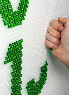 someone is stitching the letters on a pillow with green beads and thread, while another person holds a pen in their left hand