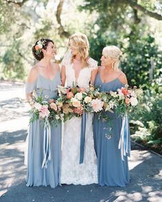 three bridesmaids in blue dresses holding bouquets and smiling at each other with trees in the background