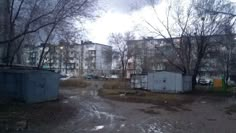 an abandoned building in the middle of a dirty lot with trees and buildings behind it