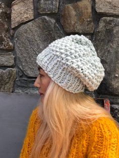 a woman with blonde hair wearing a white knitted hat next to a stone wall