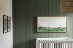 a white crib in front of a green wall with pictures on it and a painting hanging above