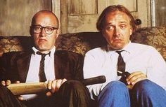 two men sitting on a couch one holding a baseball bat and the other wearing glasses