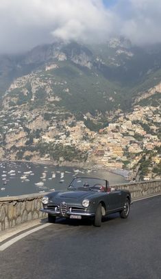 an old car is driving down the road by the water and mountains in the background