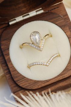 two rings in a wooden box with white feathers on the floor behind them and one ring has a pear shaped diamond