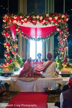 the bride and groom are sitting in front of an arch decorated with red, white and pink flowers