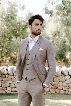 a man wearing a suit and bow tie standing in front of a stone wall with trees