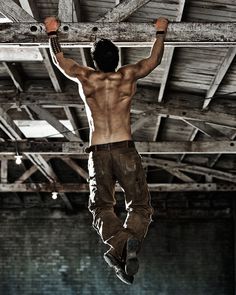 a shirtless man hanging upside down on a wooden beam in an old building with exposed beams