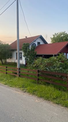 a house with a red roof is next to a wooden fence and green grass on the side of the road