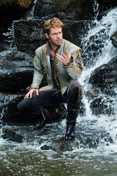 a man dressed in medieval clothing sitting on rocks next to a waterfall and holding his hand out