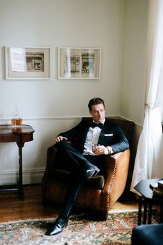 a man in a tuxedo is sitting on a chair and holding a drink