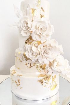 a white and gold wedding cake with flowers on it's tiers, sitting on top of a glass plate