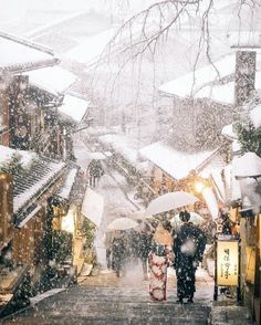 two people walking down a snowy street holding umbrellas in the snow with buildings and trees behind them