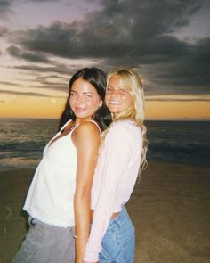 two beautiful young women standing next to each other on a beach at sunset with the ocean in the background