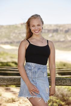 a woman standing next to a wooden fence wearing a black tank top and denim skirt