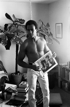 a shirtless man standing in a living room holding a newspaper