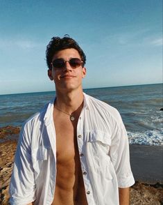 a shirtless man standing on the beach with his hands in his pockets and wearing sunglasses