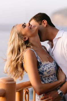 a man kissing a woman on the cheek while standing next to a railing by the ocean