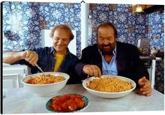 two men sitting at a table with bowls of spaghetti in front of them, one holding a fork and the other eating