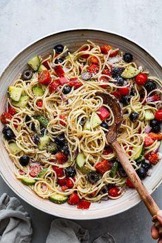 a bowl filled with pasta and vegetables on top of a white table cloth next to a wooden spoon