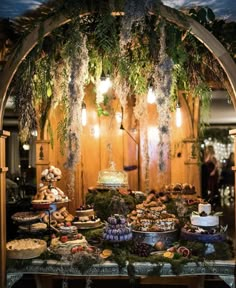 a table topped with lots of food under a wooden arch covered in greenery and hanging lights