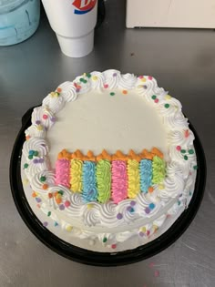 a birthday cake with white frosting and multi colored icing on the top is sitting on a table next to a cup