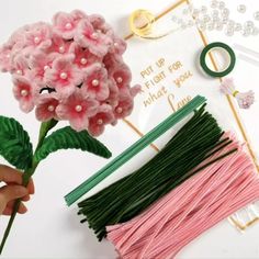 a person holding a pink flower next to some knitting needles and beads on a table