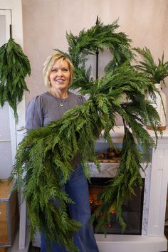 a woman is holding a wreath made out of green leaves and branches in front of a fireplace