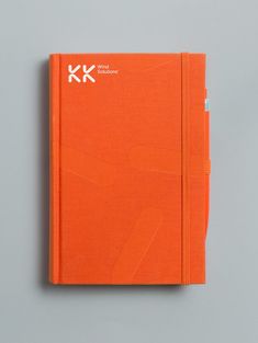 an orange notebook sitting on top of a gray surface with the letter k in white