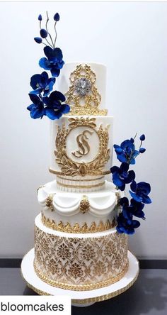 a white and gold wedding cake with blue flowers on the top tier is decorated with monograms