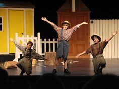 three people are performing on stage in front of a white picket fence and yellow building