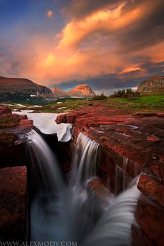 a waterfall in the middle of a rocky area with mountains in the background at sunset