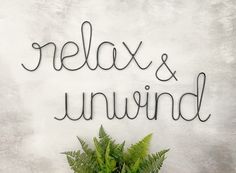 the words relax and unwind written in cursive writing on a white wall next to a potted plant