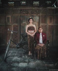 a man and woman standing next to each other in front of a wooden door with an old bicycle