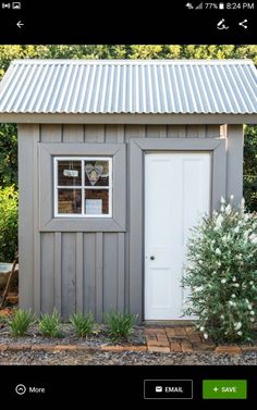 a small gray shed with a white door and window on the side, in front of some bushes