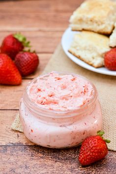 a jar of strawberry butter next to some strawberries on a wooden table with biscuits and biscuits in the background