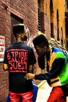 two men standing next to each other in front of a brick wall with graffiti on it