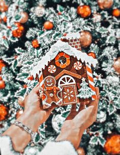 two hands holding a gingerbread house in front of a christmas tree with orange ornaments