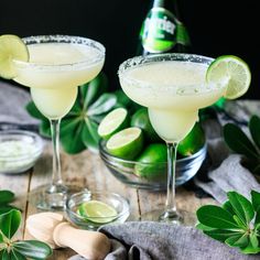 two margaritas sitting next to each other on a table with limes in the background